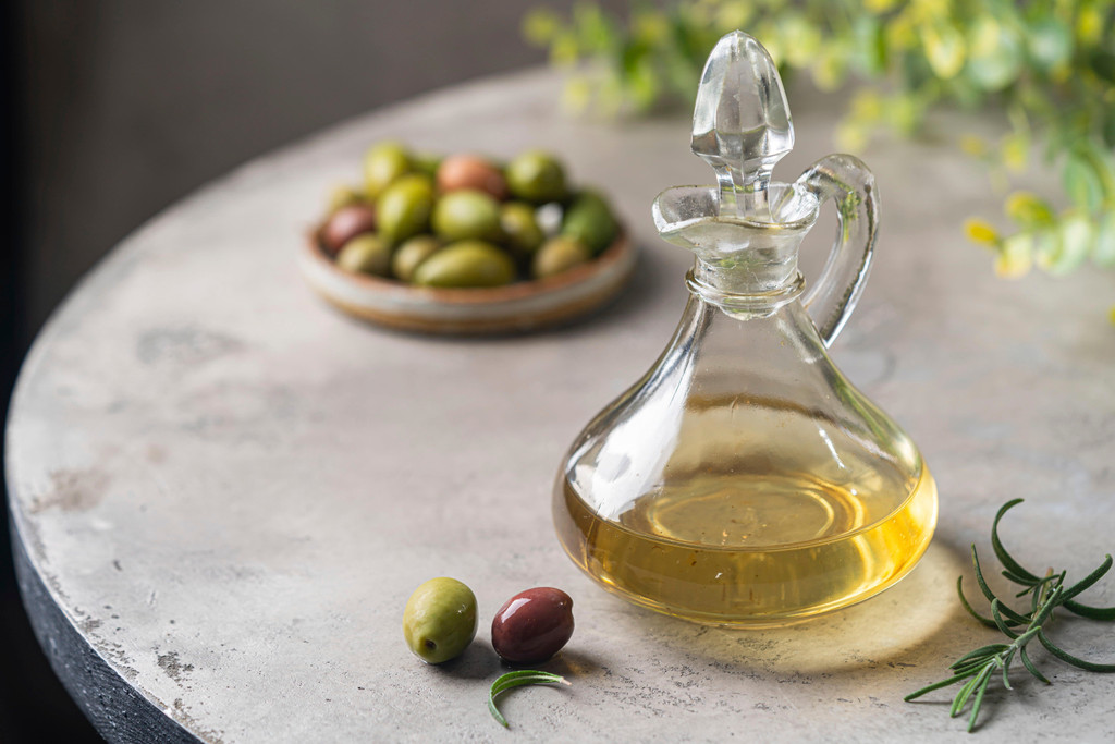 Olivenöl in einem Glasflaschenbehälter und Oliven mit Kräutern auf grauem Hintergrund ernten.