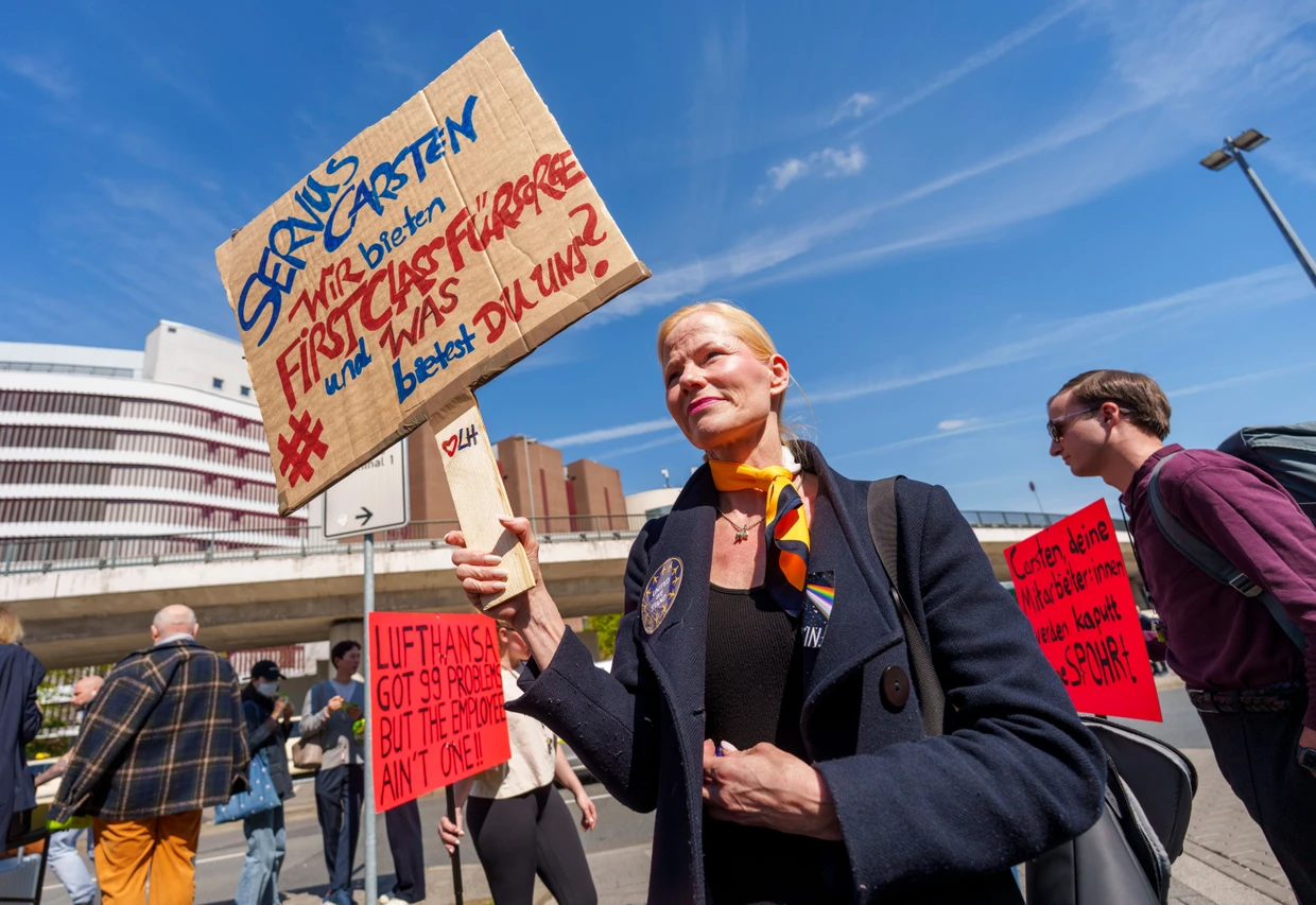 Eine Flugbegleiterin hält ein Schild "Servus Carsten Wir bieten First Class Fürsorge und was bietest Du uns?" hoch.