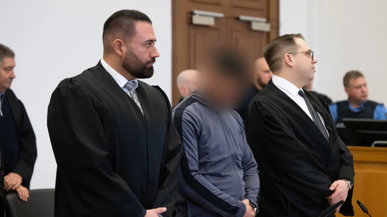 Einer der Angeklagten steht im Gerichtssaal zwischen seinen Anwälten.