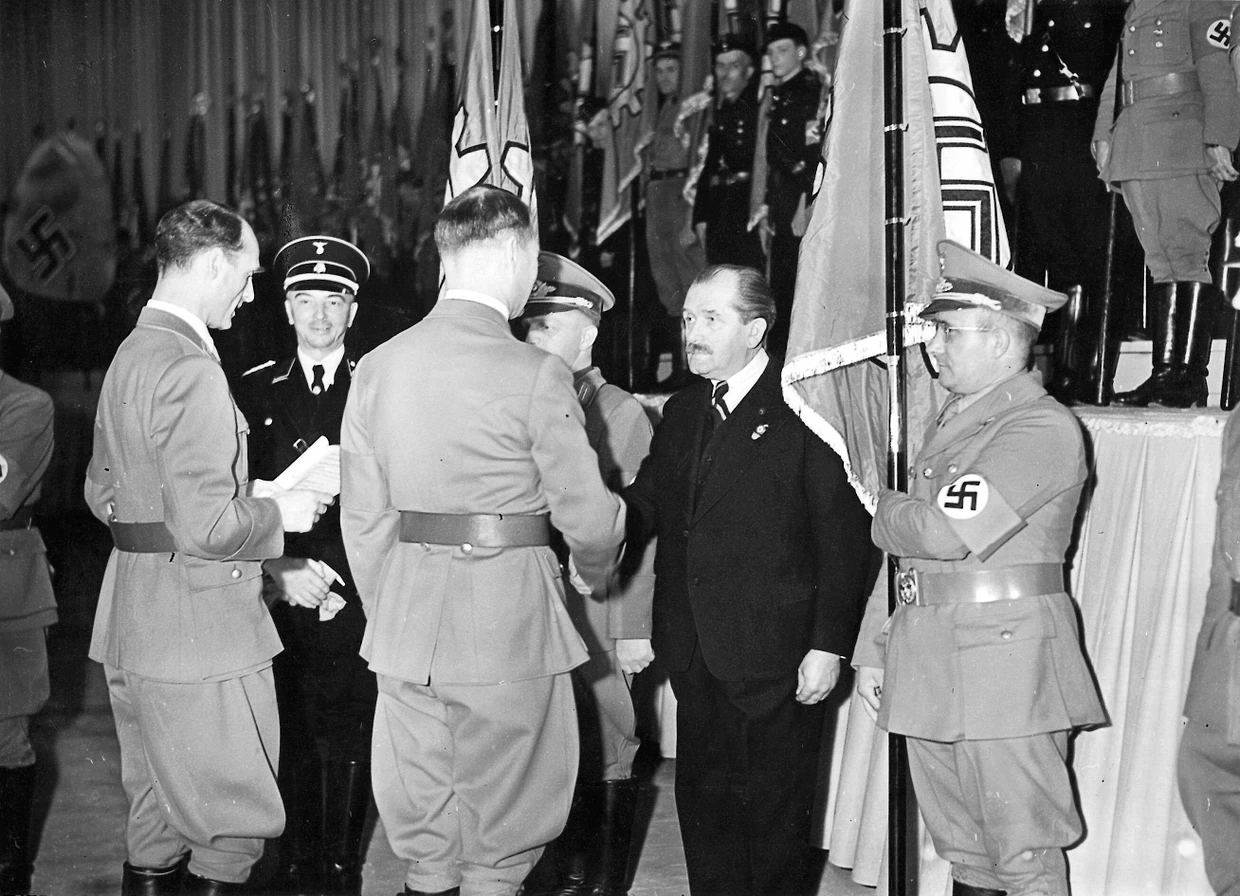 Bei der Tagung der Reichsarbeitskammer in Augsburg zeichnet Rudolf Heß in einer Halle der Messerschmittwerke Ferdinand Porsches Betrieb als nationalsozialistischen Musterbetrieb aus.