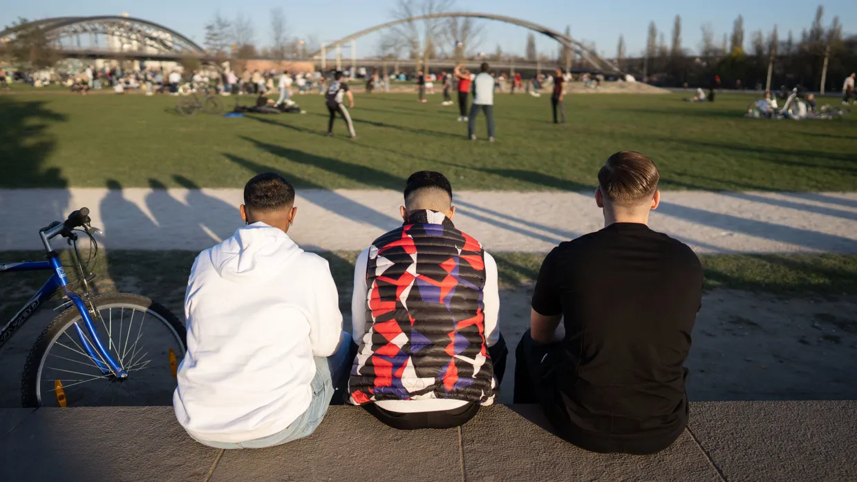 Drei junge Männer sitzen bei sonnigem Wetter auf einer Mauer im Hafenpark. (Archivbild)