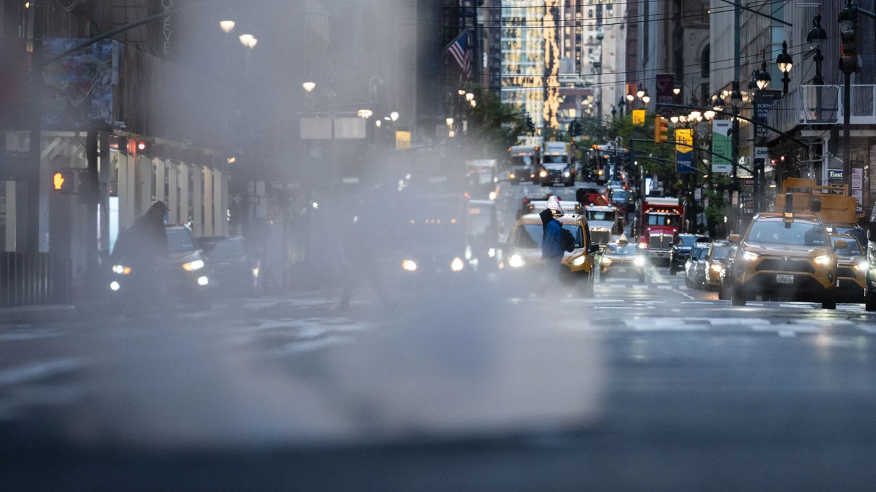 Verkehr ist am frühen Morgen im Stadtteil Manhattan unterwegs, aus einem Gully steigt Dampf auf.