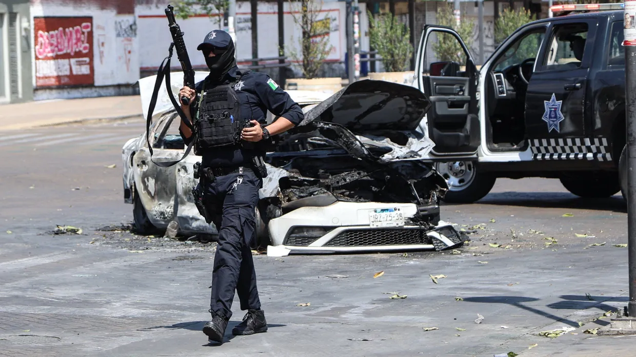 Gewalt nach „El Mencho“s Tod: Ein Polizist steht in Guadalajara neben einem verbrannten Auto auf der Straße Wache.
