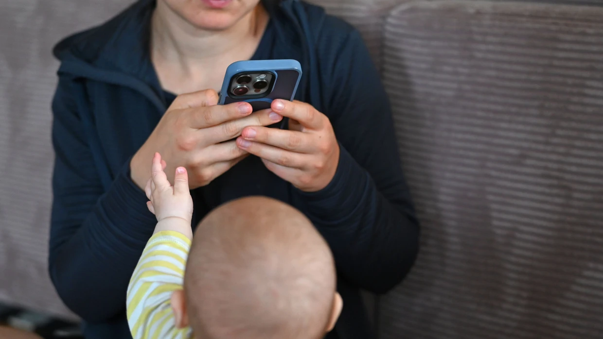 A baby reaches for his mother's smartphone while she looks at the display.