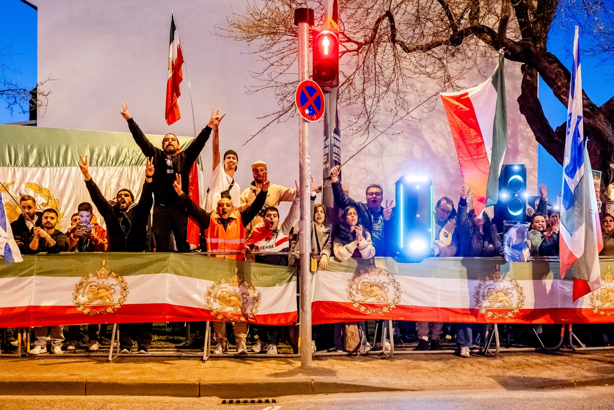 Die Gegendemonstranten verschaffen sich mit Rufen und Musik Gehör. Sie feiern die Angriffe der USA und Israels auf Iran und sind an den Flaggen mit dem Löwensymbol als Anhänger des Schah-Sohns Reza Pahlavi zu erkennen.