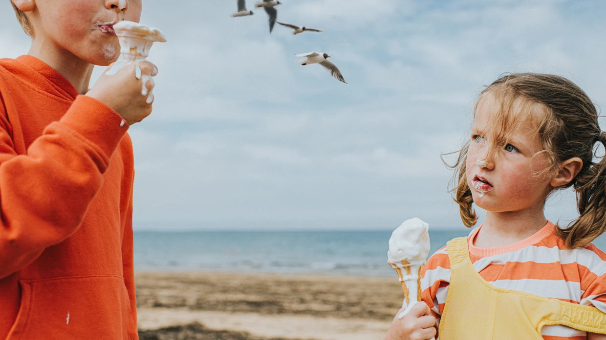 Zwei Kinder, ein Junge und ein Mädchen, stehen an einem Strand. In den Händen halten sie ein Eis, das bereits schmilzt.