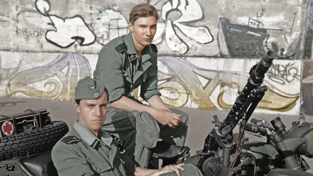 Dieses Foto stammt aus dem August 2022. Es zeigt Freiwillige, die auf der polnischen  Halbinsel Hel die Eroberung der Normandie durch alliierte Truppen im Sommer 1944 nachstellen. Die Fotografin Natalia Kepesz hat es in Schwarz-Weiß auf Film fotografiert und nachträglich eingefärbt, als wäre es eine historische Aufnahme. Im Hintergrund ist modernes Graffiti zu sehen.