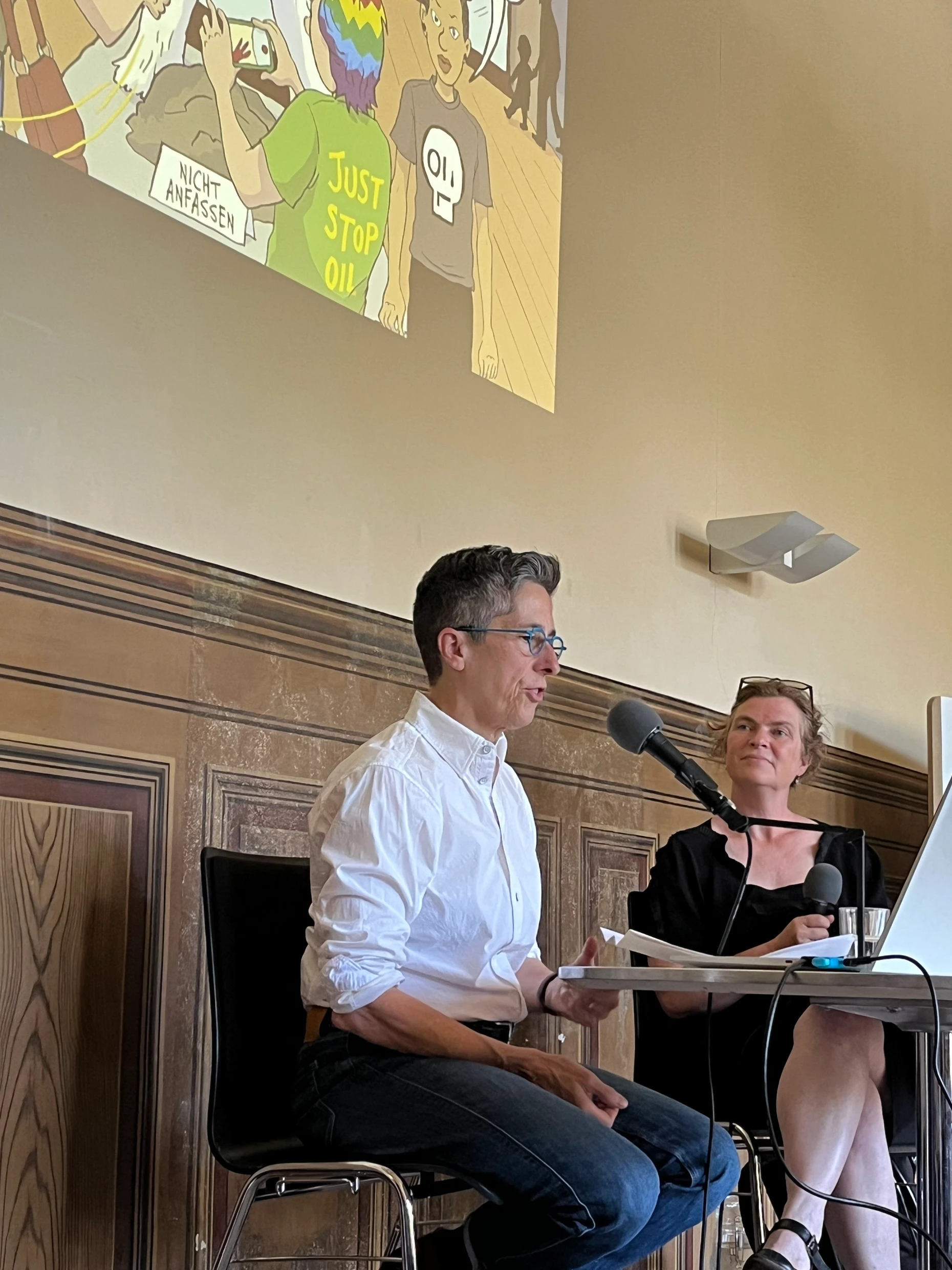 Η Alison Bechdel με την Gesa Ufer στο Salon Graphic Literature του Βερολίνου. Η σκηνή από το «Kaputt» που προβάλλεται στον τοίχο δείχνει μια επίθεση με μπογιά από νεαρούς ακτιβιστές για το κλίμα στο Μουσείο Φυσικής Ιστορίας του Βερμόντ.