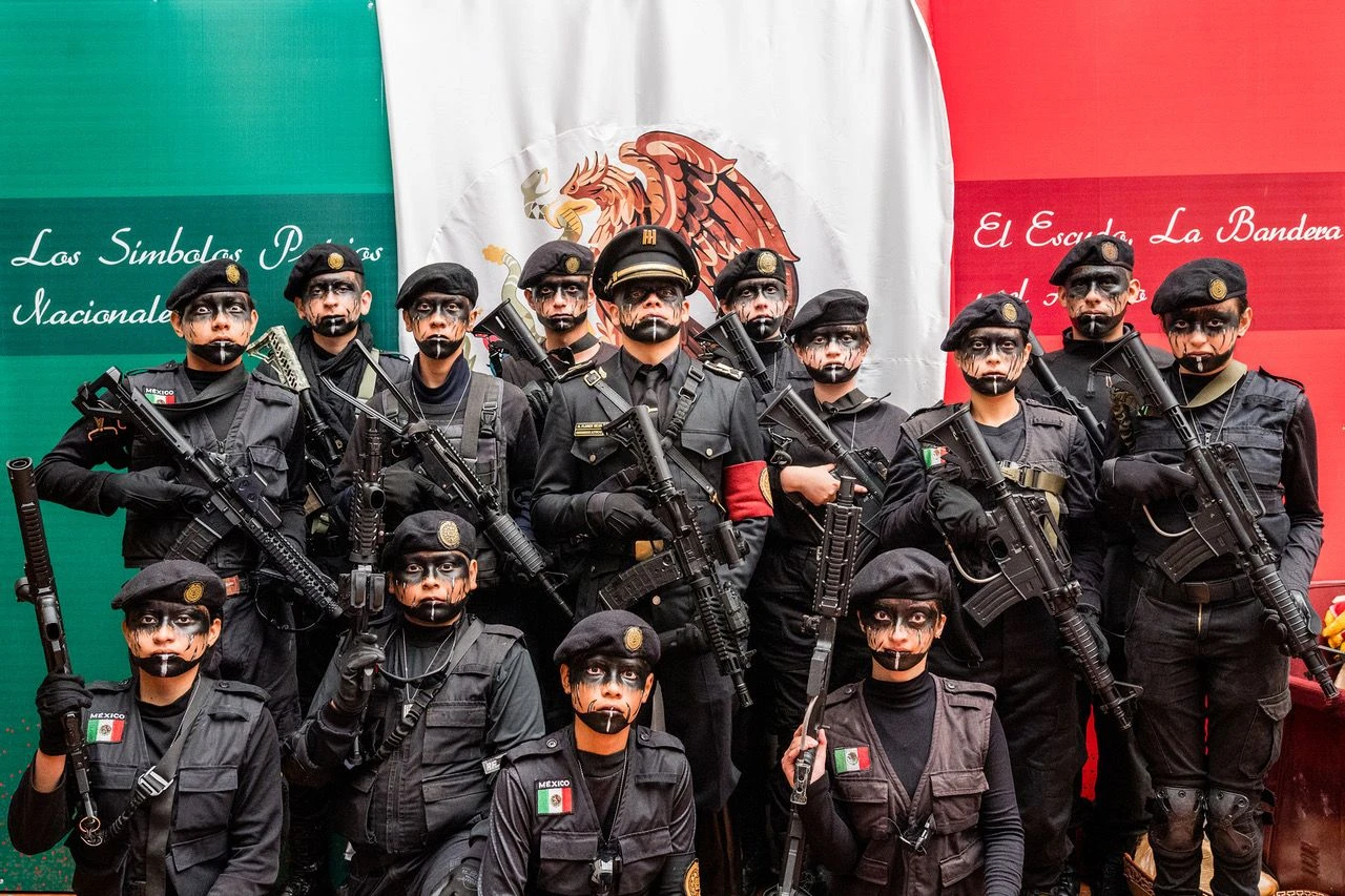 Uniformierte Kinder mit Gewehren und ihr Kommandant posieren vor der mexikanischen Flagge. In einem chaotischen Alltag steht das Militär für Verlässlichkeit und Struktur.