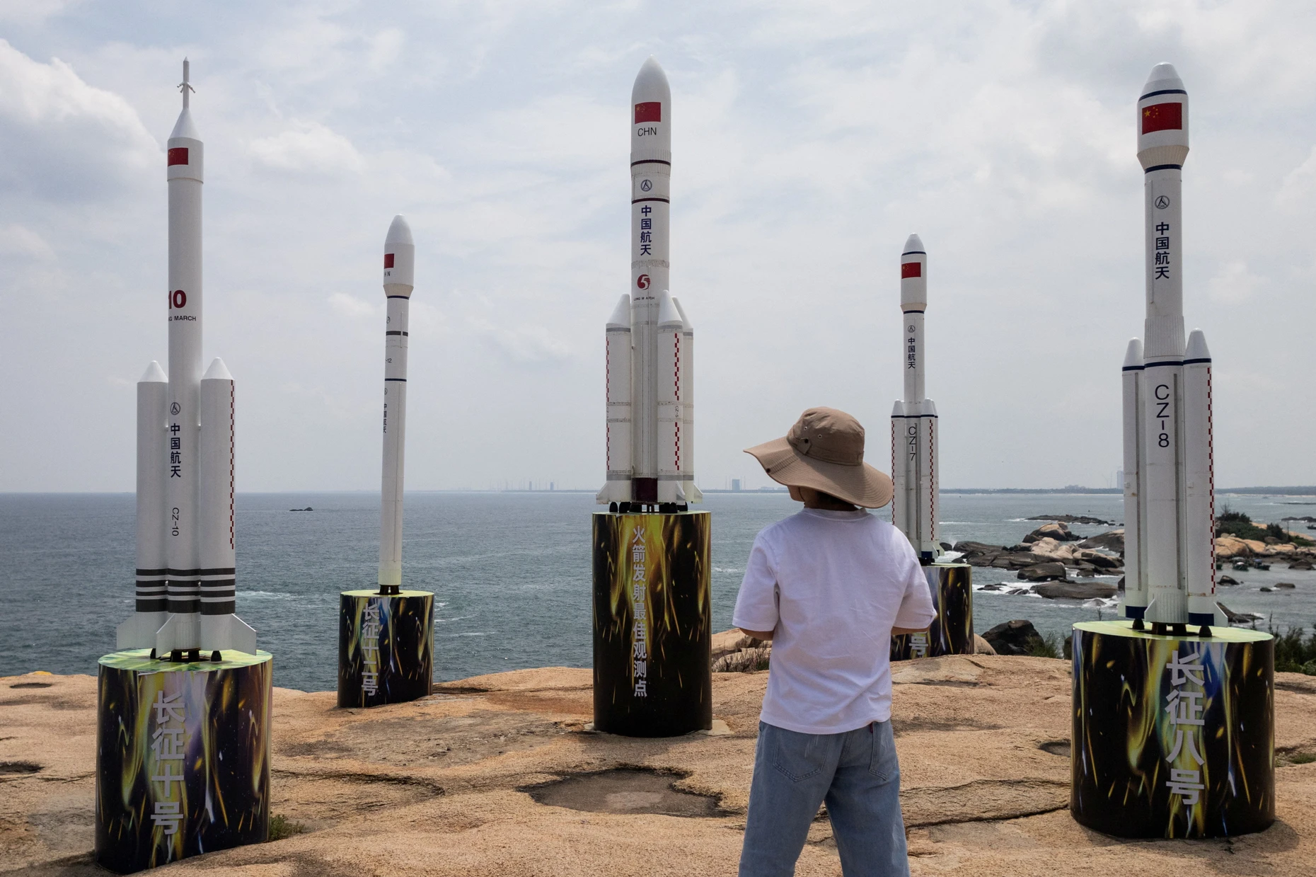 Ein Mann geht an Modellen chinesischer Trägerraketen vorbei. In der Ferne ist das Raumfahrtzentrum Wenchang zu sehen. Wenchang Rock Park.