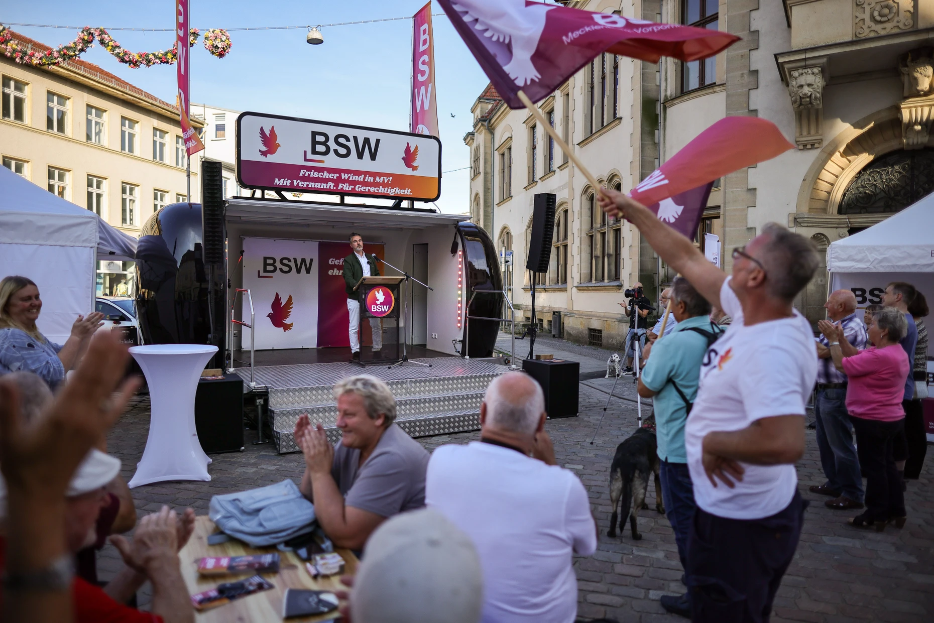 Warnt vor „verkümmerten Innenstädten“: BSW-Anhänger schwenken während De Masis Rede in Güstrow Fahnen der Partei.