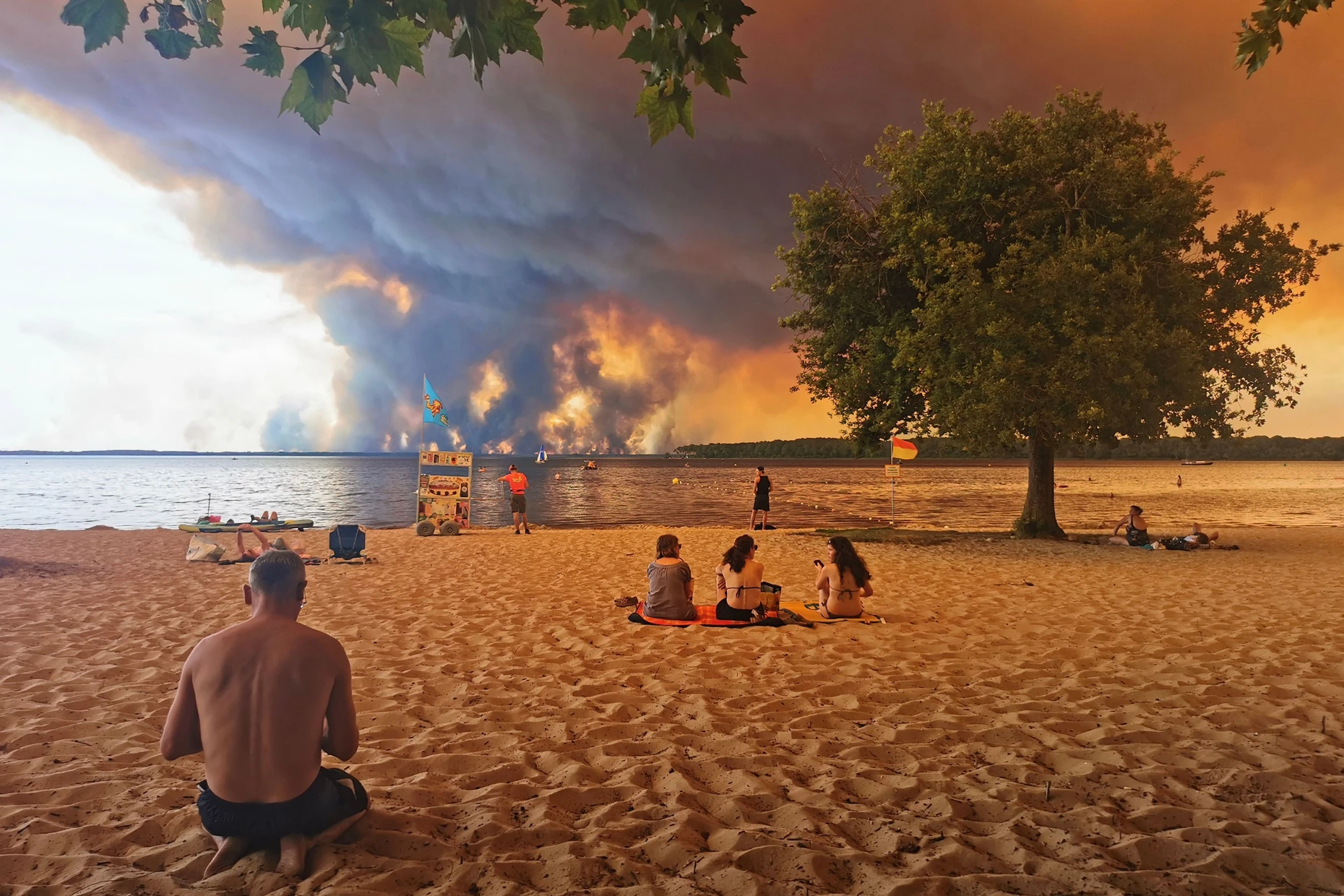 Strandbesucher sitzen am Strand von Moutchic in Lacanau im Südwesten Frankreichs, während Rauchwolken von einem Waldbrand in den Himmel aufsteigen.