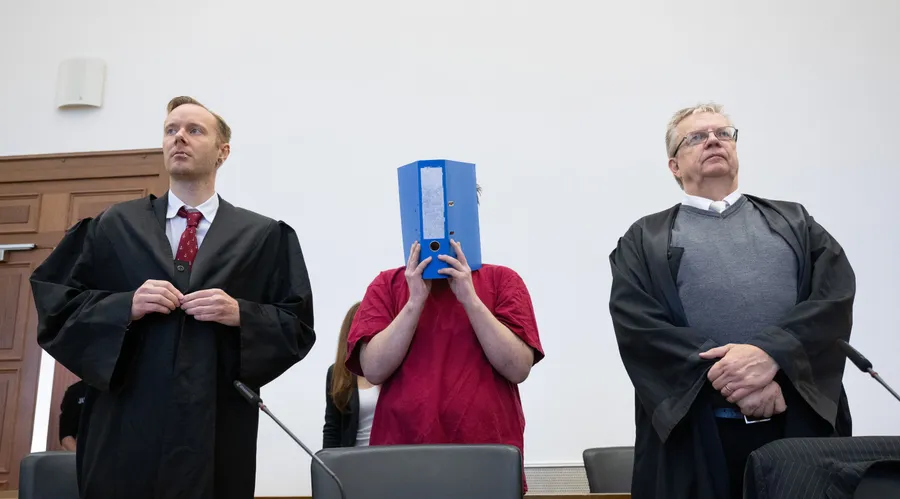 Der Angeklagte im Mordfall der Schülerin Ayleen wird in den Verhandlungssaal geführt und verbirgt sein Gesicht hinter einem Aktenordner.