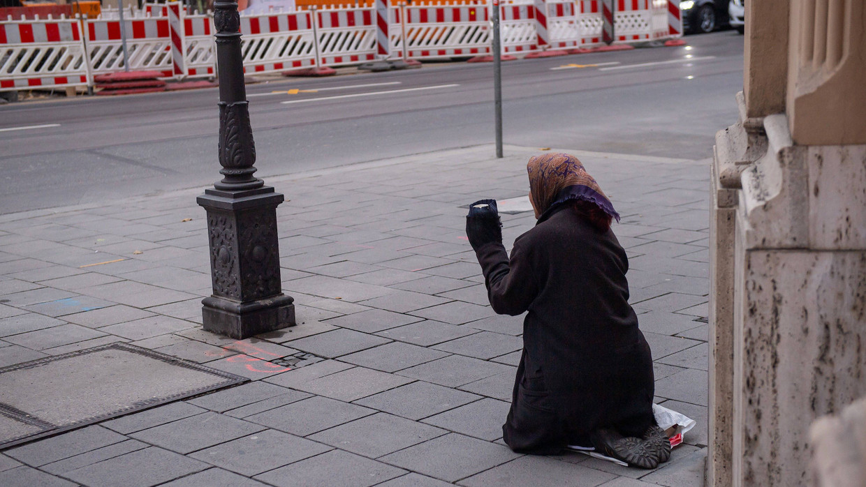 Eine bettelnde Frau kniet auf dem Gehweg einer Einkaufsstraße.