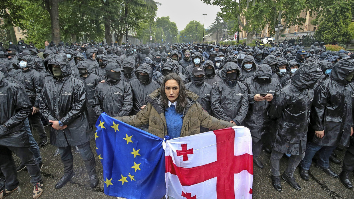 Eine Frau mit der georgischen und der EU-Fahne steht vor Polizisten, die eine Straße sperren, um Oppositionelle aufzuhalten.